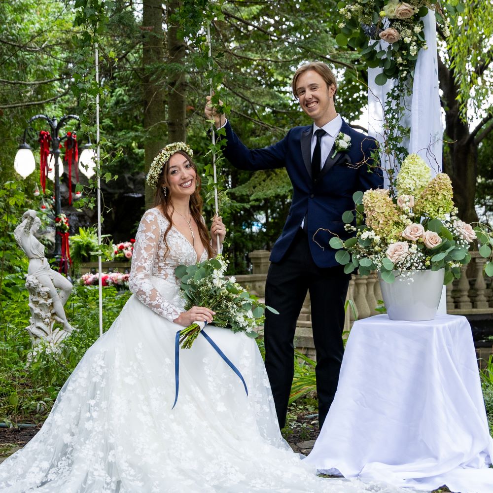 Couple de mariés souriant sous arche florale bohème, jardinière
et bouquet visibles, moment de joie mariage poétique blanc