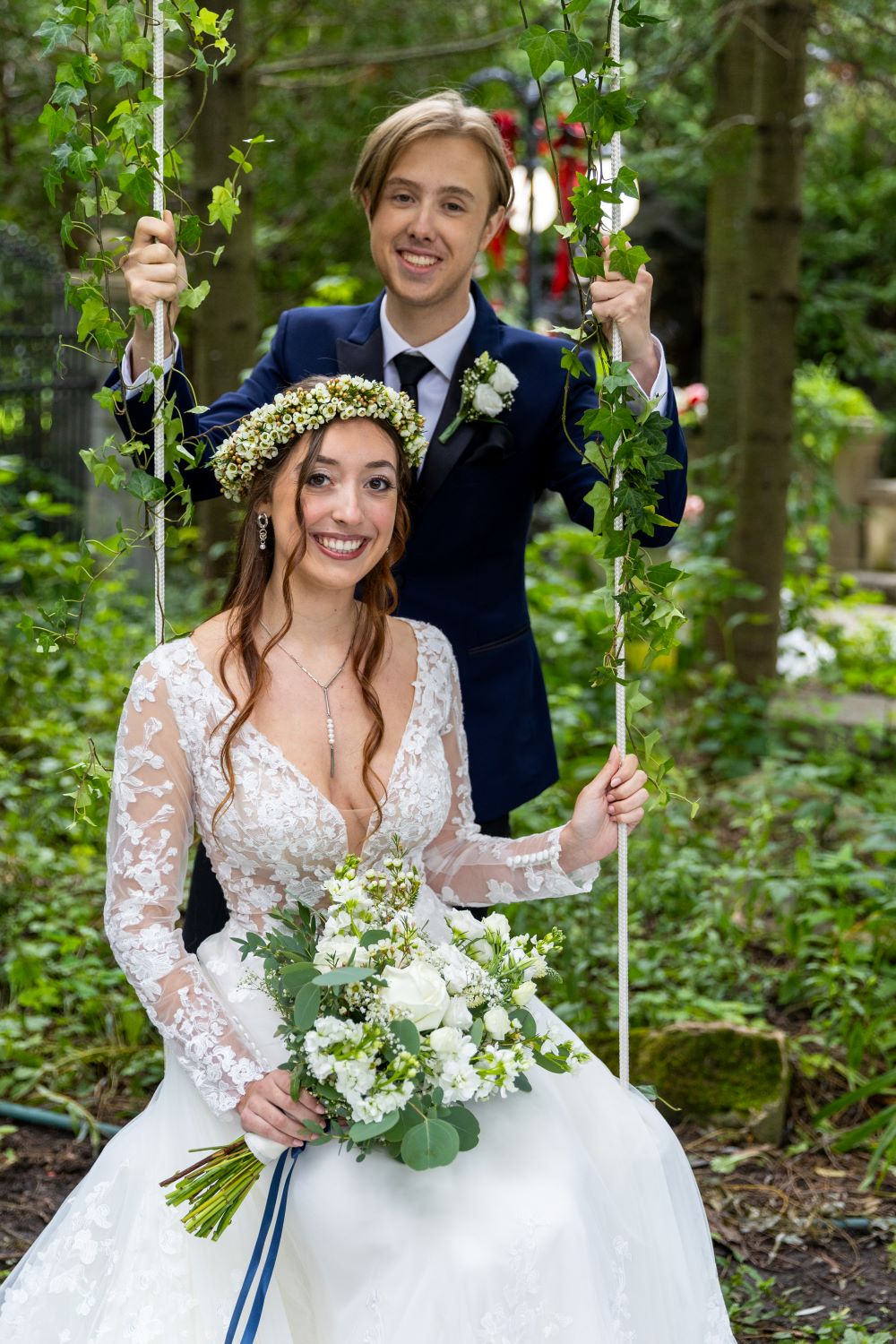 Couple de mariés, mariée assise sur balançoire avec couronne de fleurs wax,
marié debout derrière elle, moment romantique mariage bohème